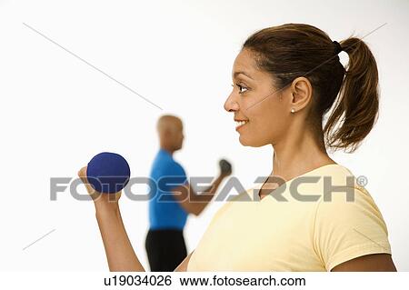 Stock Photograph - Close up of smiling mid adult woman exercising using dumbbells with mid adult man in background.. Fotosearch