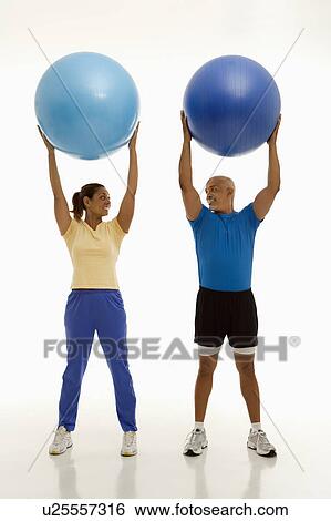 Stock Photograph - Mid adult man and woman holding blue exercise balls over their heads looking at each other smiling. Fotosearch