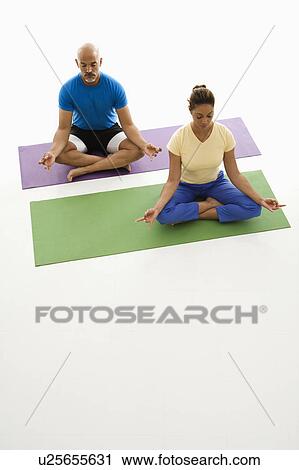 Stock Image - Mid adult man and woman sitting in lotus position on exercise mats with eyes closed and legs crossed. Fotosearch