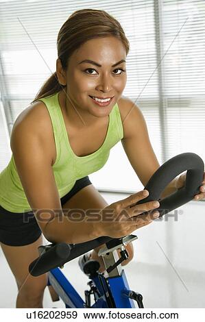 Stock Photo - Mid adult woman pedaling exercise bicycle smiling at viewer.. Fotosearch