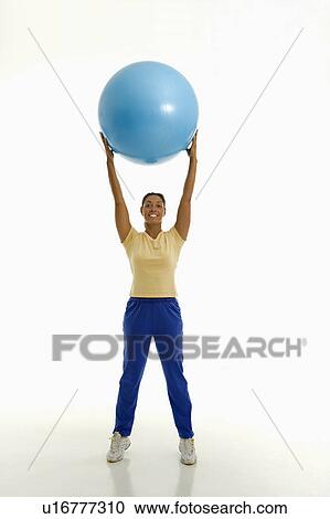 Stock Image - Mid adult woman standing and holding blue exercise ball over her head looking at viewer and smiling.. Fotosearch