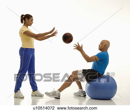 Stock Image - Mid adult woman throwing ball to mid adult man balancing on blue exercise ball. Fotosearch