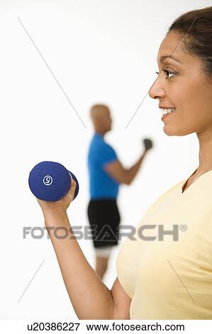 Woman exercising using dumbbells with man in background View Large Photo Image Stock Photo - Woman exercising using dumbbells with man in background. Fotosearch