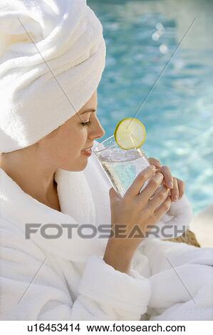 Picture - Mid-adult woman wearing robe and towel on head drinking from glass next to pool. Fotosearch