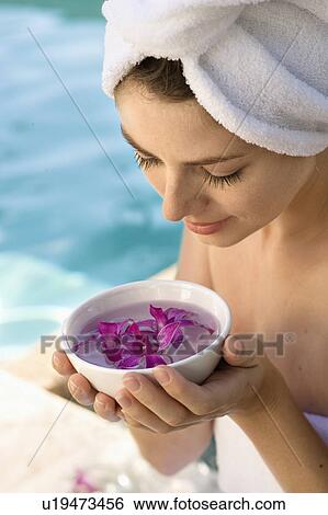 Stock Photograph - Woman wearing towel around head and body holding bowl of purple orchids next to pool. Fotosearch