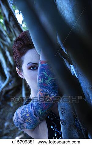 Blue-toned portrait of tattooed Caucasian woman hiding behind Banyan tree View Large Photo Image Stock Image - Blue-toned portrait of tattooed Caucasian woman hiding behind Banyan tree . Fotosearch
