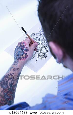 Stock Image - Caucasian male tattoo artist drawing tattoo on light table.. Fotosearch