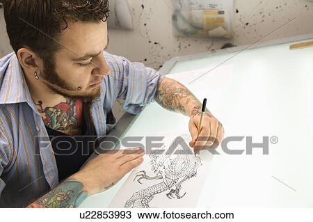 Stock Image - Caucasian male tattoo artist drawing tattoo on light table.. Fotosearch