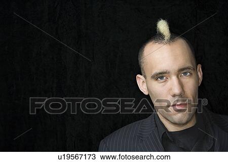Stock Image - Portrait of Caucasian man in suit with mohawk against black background.. Fotosearch