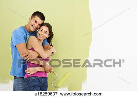 Stock Photo - Attractive smiling couple posing in front of partially painted wall holding paint roller.. Fotosearch