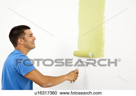 Stock Image - Attractive smiling man painting white wall with green paint.. Fotosearch
