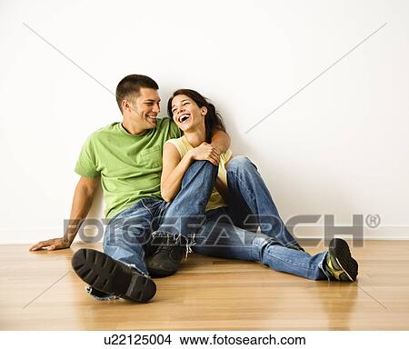Picture - Attractive young adult couple sitting close on hardwood floor in home smiling and laughing.. Fotosearch