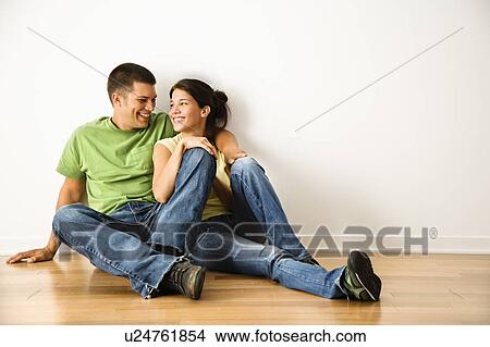 Picture - Attractive young adult couple sitting close on hardwood floor in home smiling. Fotosearch