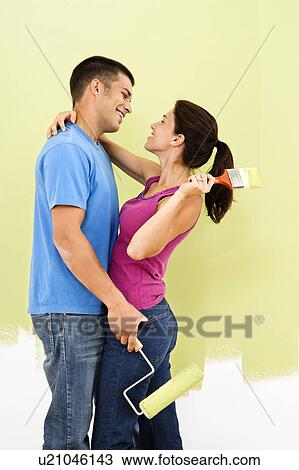 Couple hugging and smiling at each other holding paintbrushes in front of partially painted wall. View Large Photo Image Stock Image - Couple hugging and smiling at each other holding paintbrushes in front of partially painted wall.. Fotosearch