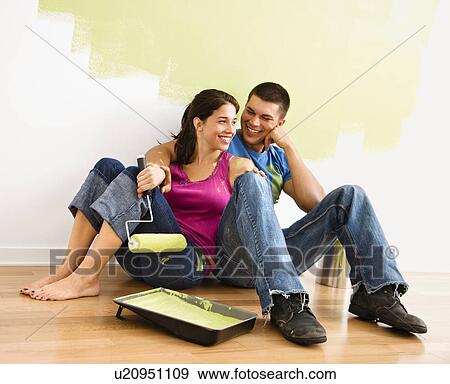 Stock Photo - Couple sitting on floor smiling in front of partially painted wall in home.. Fotosearch