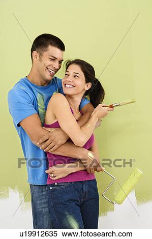 Couple smiling and embracing in front of partially painted interior wall holding paintbrush and roller. View Large Photo Image Stock Photography - Couple smiling and embracing in front of partially painted interior wall holding paintbrush and roller.. Fotosearch
