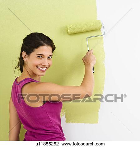 Stock Image - Pretty smiling woman painting interior wall of home with paint roller.. Fotosearch