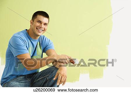 Stock Photo - Smiling man kneeling in front of partially painted wall holding paintbrush. Fotosearch