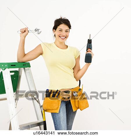 Stock Image - Woman standing in home with ladder and holding tools smiling.. Fotosearch