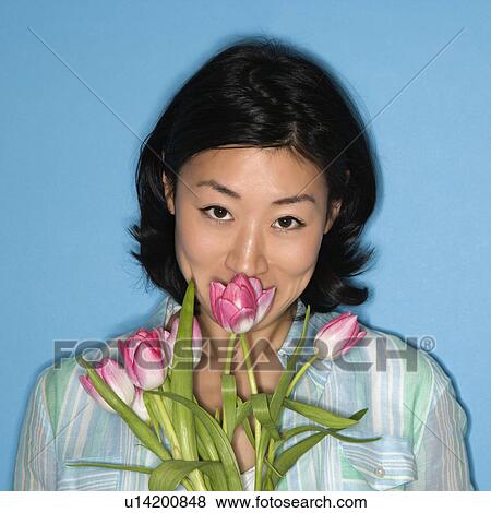 Stock Photo - Head and shoulder portrait of pretty woman smelling flowers on blue background. Fotosearch