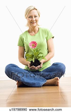 Stock Image - Portrait of smiling adult blonde woman sitting on floor holding pink gerbera daisy plant.. Fotosearch