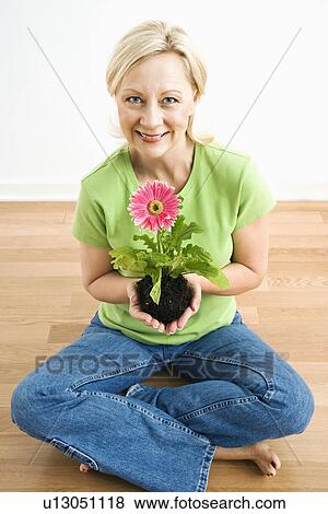Stock Photo - Portrait of smiling adult blonde woman sitting on floor holding pink gerbera daisy plant.. Fotosearch