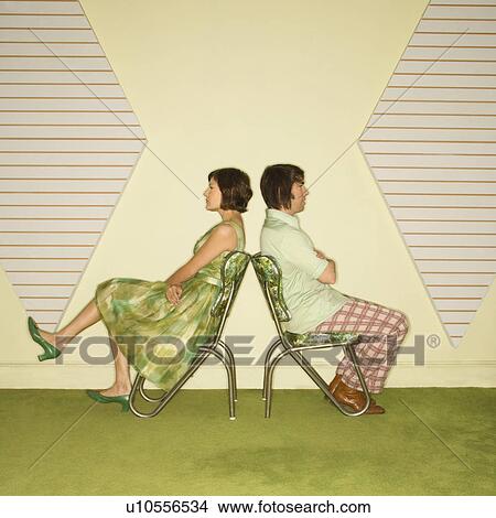 Picture - Caucasian mid-adult couple wearing vintage clothing sitting back to back in green vinyl chairs with arms crossed.. Fotosearch
