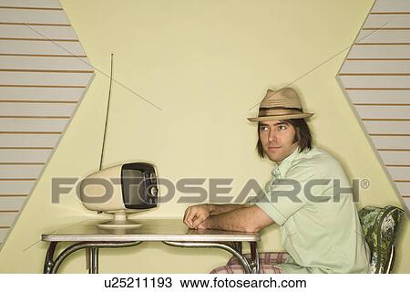 Stock Image - Caucasian mid-adult man wearing hat sitting at 50's retro dinette set in front of old television.. Fotosearch