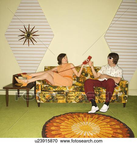 Stock Image - Caucasian mid-adult woman passing phone to Caucasian mid-adult man wearing sunglasses sitting on sofa.. Fotosearch
