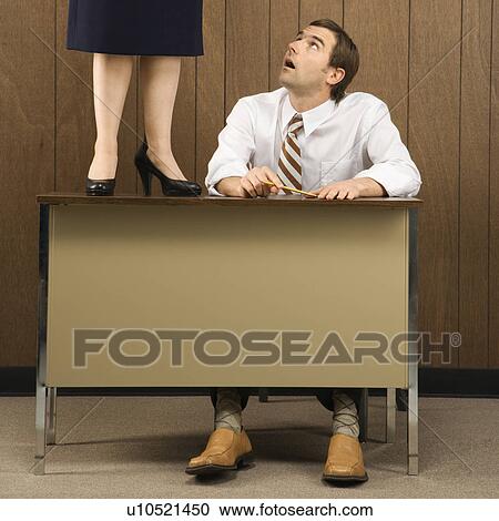 Stock Image - Mid-adult Caucasian male sitting at desk looking up to Caucasian female standing on desk.. Fotosearch