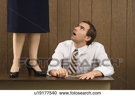 Stock Image - Mid-adult Caucasian male sitting at desk looking up to Caucasian female standing on desk.. Fotosearch