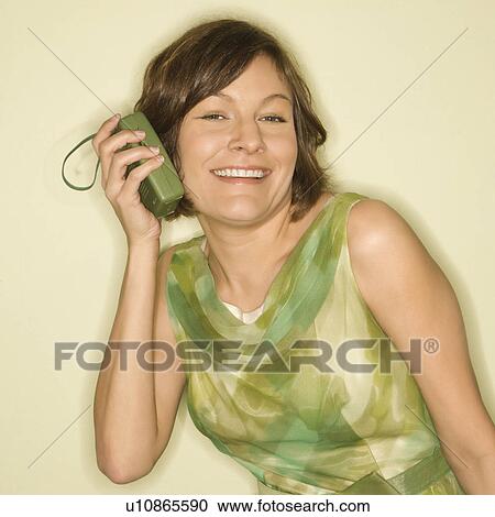 Stock Image - Pretty Caucasian mid-adult woman wearing green vintage dress holding handheld radio up to her ear and smiling at viewer.. Fotosearch