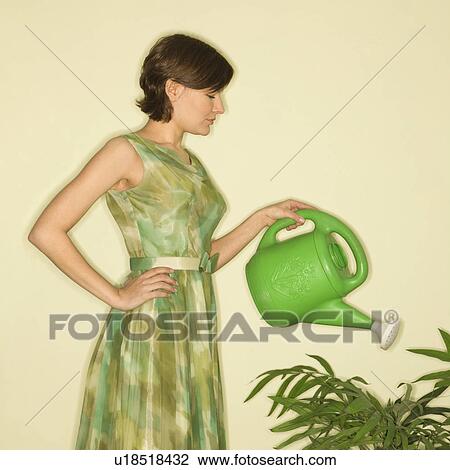 Stock Image - Pretty Caucasian mid-adult woman wearing vintage dress watering houseplant with green watering can.. Fotosearch
