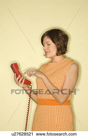 Stock Image - Pretty Caucasian mid-adult woman wearing orange dress standing dialing red telephone.. Fotosearch