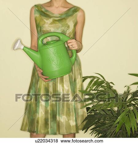 Stock Photo - Pretty Caucasian mid-adult woman wearing vintage dress standing next to houseplant holding green watering can.. Fotosearch