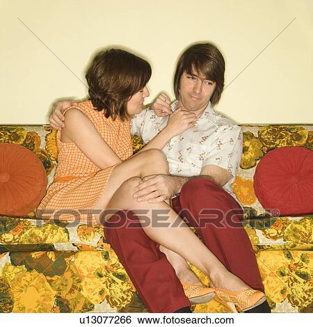 Stock Photograph - Pretty Caucasian mid-adult woman flirting with shy Caucasian mid-adult man sitting on colorful retro sofa.. Fotosearch