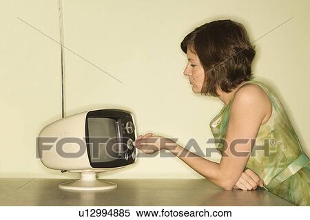 Stock Photography - Side view of pretty Caucasian mid-adult woman sitting at 50's retro dinette set turning old televsion knob.. Fotosearch