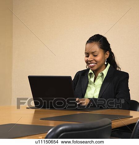 Picture - Businesswoman sitting at conference table smiling and typing on laptop computer. Fotosearch