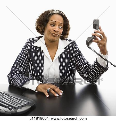Picture - Businesswoman sitting at desk holding telephone receiver out away from head looking with strange expression.. Fotosearch