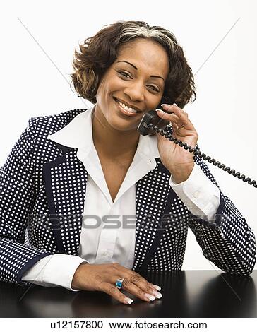Stock Image - Businesswoman sitting at desk holding telephone receiver to ear smiling. Fotosearch