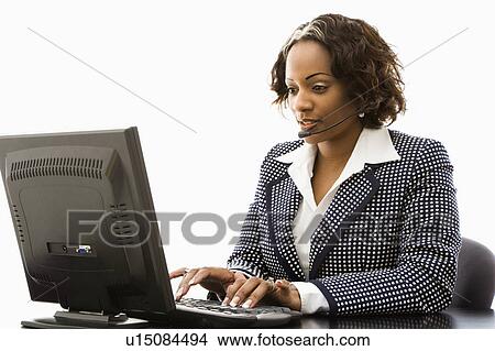 Businesswoman sitting at office desk typing on computer and talking into telephone headset. View Large Photo Image Picture - Businesswoman sitting at office desk typing on computer and talking into telephone headset.. Fotosearch