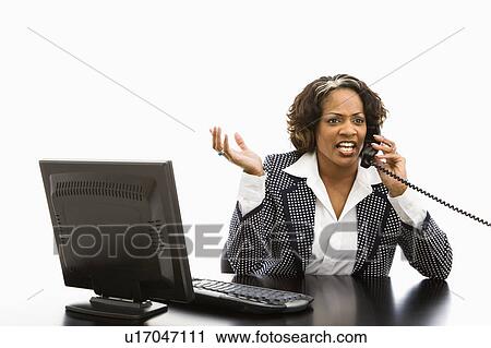 Stock Image - Businesswoman sitting at office desk with computer talking on telephone looking upset. Fotosearch