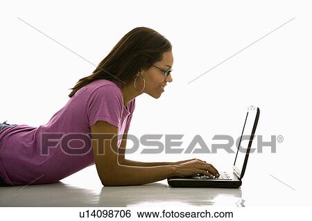 Stock Photograph - Side view of mid adult African American woman lying on floor using laptop.. Fotosearch
