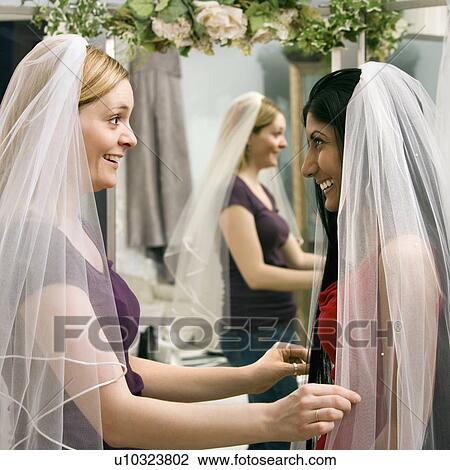 Stock Image - Indian woman and Caucasian woman trying on veils and looking in mirror.. Fotosearch