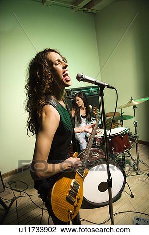 Stock Image - Caucasian females singing into microphone and playing the drums.. Fotosearch