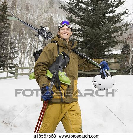 Stock Photo - Attractive man in winter clothing walking in snow carrying ski equipment and smiling.. Fotosearch