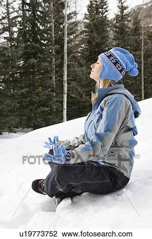 Stock Image - Side view of woman wearing blue ski clothing sitting in snow in lotus position. Fotosearch
