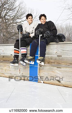 Stock Image - Two boys in ice hockey uniforms sitting on ice rink sidelines looking and smiling.. Fotosearch