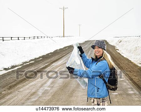 Stock Photograph - Woman in winter clothes standing on muddy dirt road holding open map smiling. Fotosearch