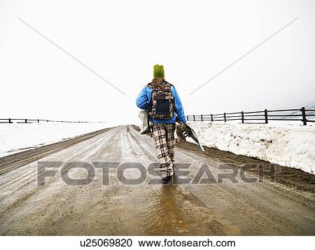 Stock Image - Young woman in winter clothes walking down muddy dirt road holding snowboard and boots.. Fotosearch